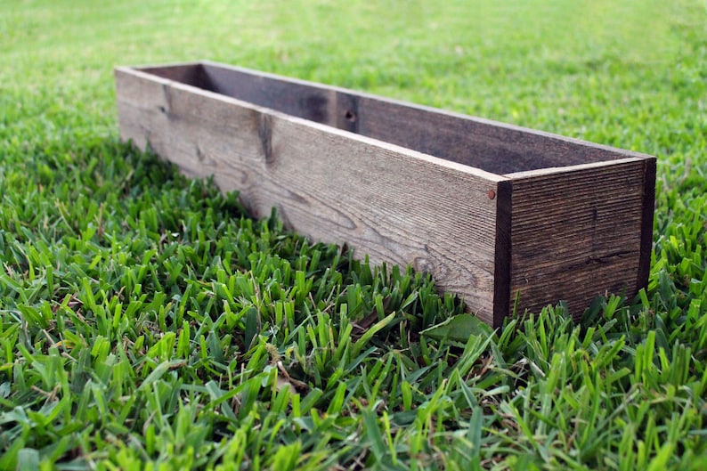 May include: A long, rectangular wooden planter box with a dark brown stain. The planter is sitting on a bed of green grass.