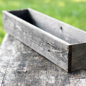 May include: A long, rectangular wooden planter box with a weathered gray finish. The box is empty and sits on a rustic wooden surface.