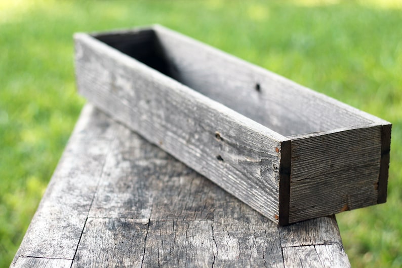 May include: A long, rectangular wooden planter box with a weathered gray finish. The box is empty and sits on a rustic wooden surface.