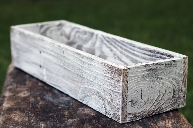 Puede incluir: Una jardinera de madera blanca con un acabado r&uacute;stico y desgastado. La jardinera es rectangular y tiene un dise&ntilde;o ancho y poco profundo.