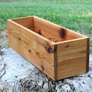 May include: A rectangular wooden planter box made of natural wood with a light brown finish. The planter is empty and sitting on a tree stump.