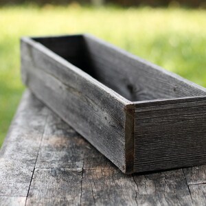 Puede incluir: Una jardinera de madera rectangular y larga, hecha de madera gris desgastada. La jardinera est&aacute; vac&iacute;a y se encuentra sobre una superficie de madera.
