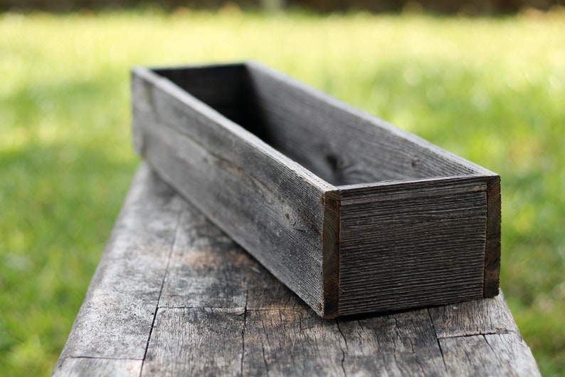 Puede incluir: Una jardinera de madera rectangular y larga, hecha de madera gris desgastada. La jardinera est&aacute; vac&iacute;a y se encuentra sobre una superficie de madera.
