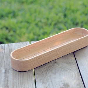 May include: A light brown wooden tray with a rounded oval shape. The tray is empty and sitting on a wooden surface.
