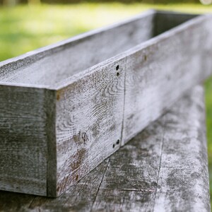 May include: A long, rectangular wooden planter box with a whitewashed finish. The planter is made of rough-hewn wood and has a rustic, farmhouse style.