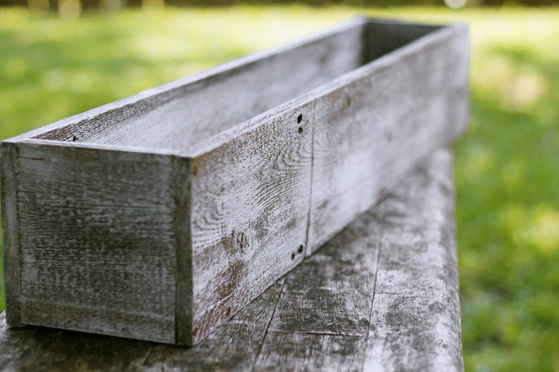 May include: A long, rectangular wooden planter box with a whitewashed finish. The planter is made of rough-hewn wood and has a rustic, farmhouse style.