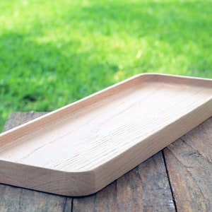 May include: A light wood rectangular serving tray with rounded edges sits on a wooden surface. The tray is empty and ready to be filled with food or other items.