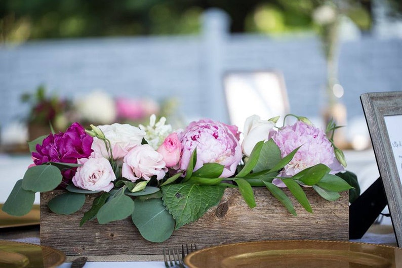 May include: A wooden box filled with pink, white, and purple flowers. The flowers are arranged in a rustic style and are surrounded by green leaves.