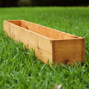 May include: A long, rectangular wooden planter box, made of natural wood, sitting on a bed of green grass.