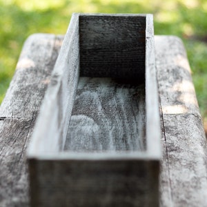 May include: A weathered wooden planter box with a rectangular shape. The box is empty and ready to be filled with plants.