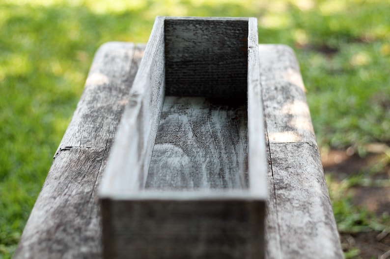 May include: A weathered wooden planter box with a rectangular shape. The box is empty and ready to be filled with plants.