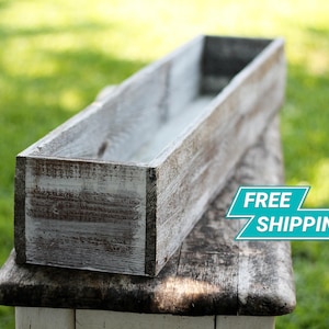 May include: A long, rectangular wooden planter box with a whitewashed finish. The planter is made of rustic wood planks and has a distressed look. The planter is sitting on a wooden surface.