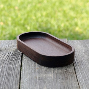 May include: A dark brown, oval-shaped wooden tray. The tray is set on a weathered, gray wooden surface, with a blurred green background. The tray's smooth interior contrasts with the rough texture of the wood.