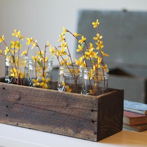 May include: A rustic wooden box planter with four glass jars filled with yellow forsythia branches. The planter is sitting on a white table.