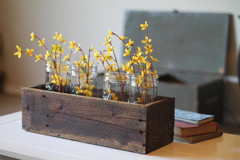 May include: A rustic wooden box planter with four glass jars filled with yellow forsythia branches. The planter is sitting on a white table.