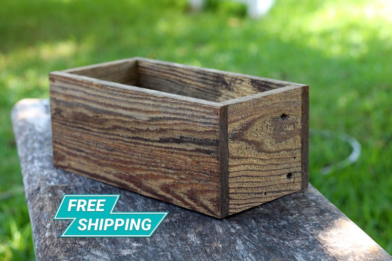 Puede incluir: Una jardinera rectangular de madera con un acabado r&uacute;stico marr&oacute;n. La jardinera est&aacute; hecha de madera reciclada y tiene un patr&oacute;n de veta de madera natural.