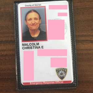 May include: A black leather badge holder with a clear plastic insert. The badge holder has a silver metal snap closure and the text "County of Warren" and "Emergency Response Official". The insert shows a photo of a woman with the text "MALCOLM CHRISTINA E" and a pink rectangular area with text that is obscured.