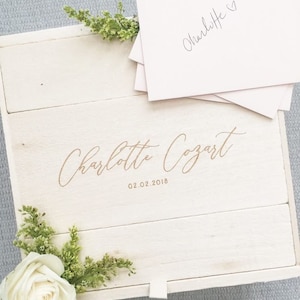 May include: A white wooden box with a gold inscription that reads "Charlotte Cozart 02.02.2018". The box is open and has a stack of white cards inside with the name "Charlotte" written on them.