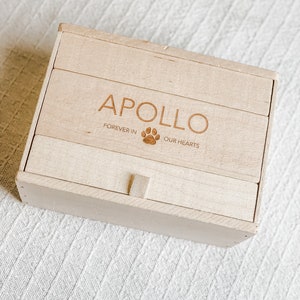 Puede incluir: Una caja de madera con tapa abatible y un peque&ntilde;o pestillo. La caja est&aacute; grabada con el nombre "Apollo" y el texto "Forever in our hearts".
