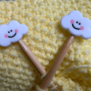 May include: Two knitting needles with white cloud-shaped toppers featuring smiling faces and pink cheeks. The needles have wooden shafts and are set against a textured yellow background, likely a knitted item.