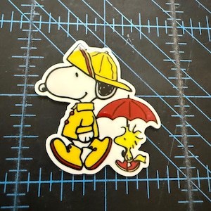 May include: A colorful sticker featuring Snoopy in a yellow raincoat and hat, holding a red umbrella with Woodstock. The cartoon characters are set against a white background with a black outline.