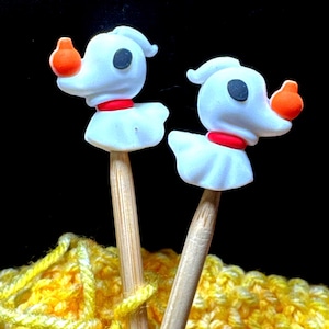 May include: Two white ghost dog knitting needles with orange pumpkins. The needles are made of wood and are resting on a yellow knitted fabric.