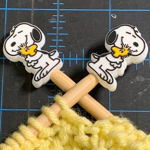 May include: Two wooden knitting needles with yellow yarn attached. Each needle has a white plastic  Snoopy character holding a yellow bone on the end.