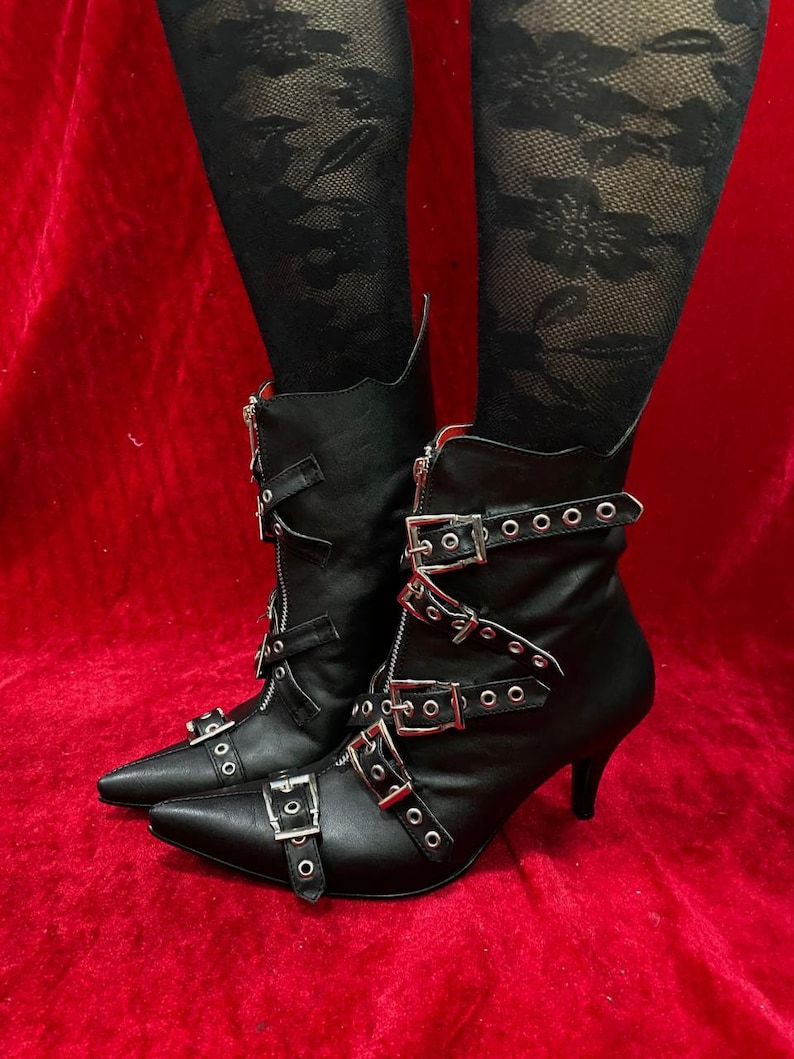 Puede incluir: Botines negros con punta, con m&uacute;ltiples hebillas y cremalleras plateadas. Los botines tienen tac&oacute;n de aguja y se llevan con medias de encaje negro, sobre un fondo rojo.