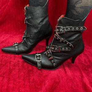 Puede incluir: Botines de cuero negro con punta y tacones de aguja. Las botas tienen hebillas plateadas, cremalleras y detalles de ojales. El interior es rojo.