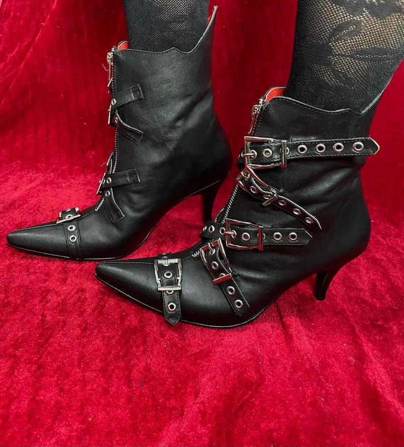 Puede incluir: Botines de cuero negro con punta y tacones de aguja. Las botas tienen hebillas plateadas, cremalleras y detalles de ojales. El interior es rojo.