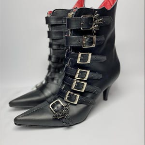 Könnte beinhalten: Ein Paar schwarze Stiefeletten mit spitzer Zehenpartie und Stilettoabsätzen. Die Stiefel haben mehrere Riemen mit silbernen Schnallen und fledermausförmigen Verzierungen. Das Innenfutter ist rot.