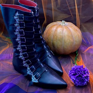 Puede incluir: Botas negras hasta la rodilla con hebillas en forma de murciélago y un interior rojo. Las botas tienen una punta afilada. Una pequeña calabaza y una flor morada están al lado de las botas.