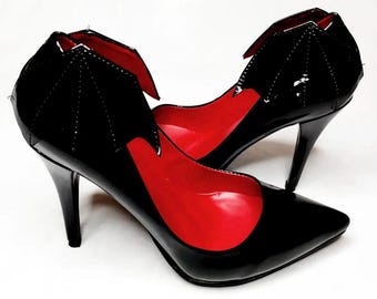 May include: Pair of black high-heeled shoes with a pointed toe and a bat wing design on the heel. The interior of the shoe is red. The shoes have a glossy finish and a stiletto heel.