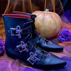 May include: Black ankle boots with silver ankh-shaped buckles and red interior lining. The boots have pointed toes and are laced up the front. A pumpkin and purple flowers are in the background.