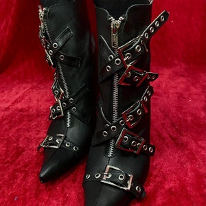 Puede incluir: Botines de cuero negro con punta, cremalleras y m&uacute;ltiples hebillas plateadas. Las botas tienen tac&oacute;n alto y est&aacute;n sobre un fondo rojo. Una elecci&oacute;n de calzado elegante y atrevida.