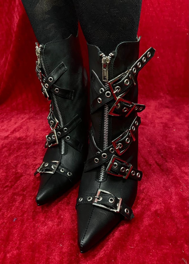 Puede incluir: Botines de cuero negro con punta, cremalleras y m&uacute;ltiples hebillas plateadas. Las botas tienen tac&oacute;n alto y est&aacute;n sobre un fondo rojo. Una elecci&oacute;n de calzado elegante y atrevida.