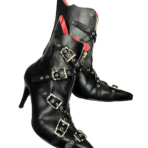 Könnte beinhalten: Schwarze Leder-Ankle Boots mit spitzer Zehenpartie, Stiletto-Absätzen und mehreren Schnallen und Riemen. Die Stiefel haben einen Reißverschluss und rotes Futter. Eine stilvolle Schuhwahl.
