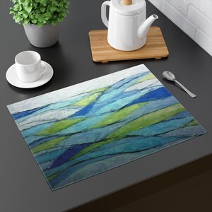 May include: A rectangular placemat with an abstract design featuring blue and green brushstrokes. The placemat is on a dark surface, with a white teacup and saucer, a teapot, and a spoon nearby.