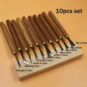 Op de afbeelding: Een set van tien houtsnijgereedschappen met houten handvatten en metalen messen. De messen hebben verschillende vormen, waaronder plat, cirkel, driehoek en schuin, met maten vari&euml;rend van 0,3 cm tot 1,5 cm. De tekst "10pcs set" is zichtbaar.