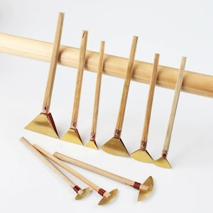 May include: A collection of handmade craft tools with wooden handles and brass blades. The tools are arranged in a row, with a larger wooden dowel in the background. The blades have a triangular or semi-circular shape.