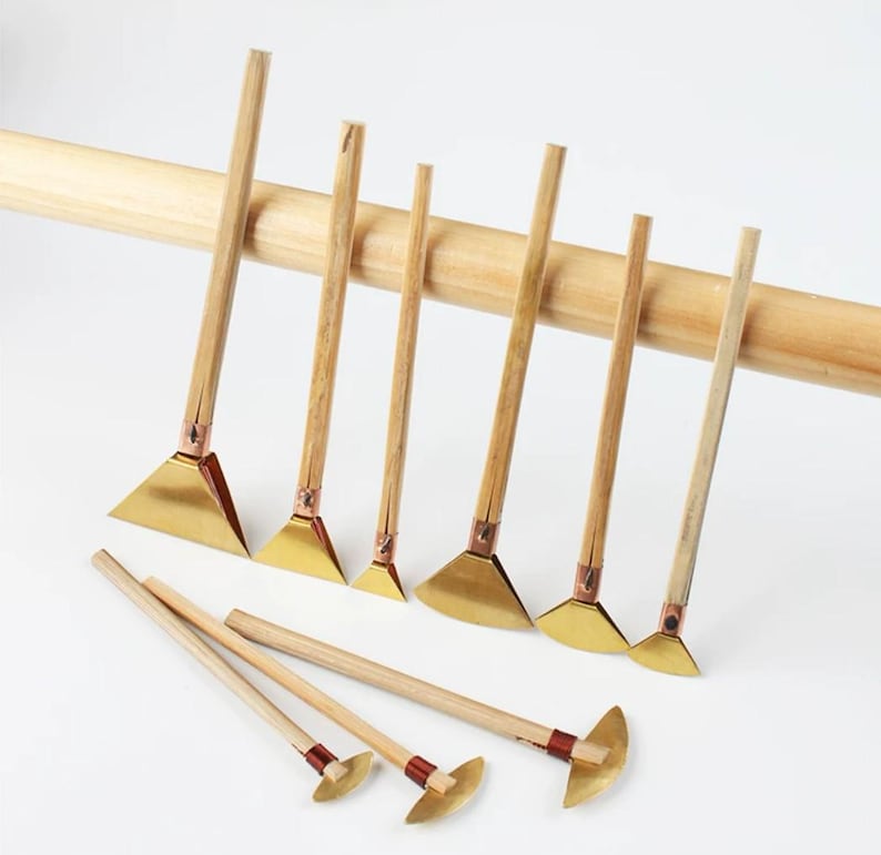 May include: A set of craft tools with wooden handles and brass blades. The tools are displayed in a row, with a larger wooden dowel behind them. The blades are either triangular or semi-circular in shape.