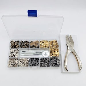 May include: A clear plastic storage box containing various metal snap fasteners in bronze, gold, silver, and black finishes, along with two metal setting tools and a circular base. A silver metal hole punch tool is positioned next to the box.