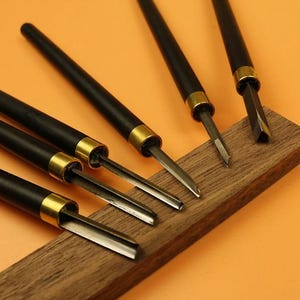 May include: A collection of wood carving tools featuring black handles and gold-toned metal ferrules. The tools are displayed on a small, rectangular wooden block. The blades are visible, and the background is a solid orange colour.