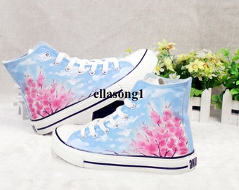 May include: A pair of high-top canvas sneakers with a hand-painted design. The sneakers feature a light blue sky background with pink cherry blossom branches. White laces and soles complete the design. The text "ellasong1" is visible.