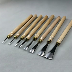 May include: A set of eight wood carving tools with wooden handles and metal blades. The blades have different shapes and sizes, with numbers 2, 4, 6, 8, 10, 15, 20, and 25 printed on them. The tools are arranged diagonally.