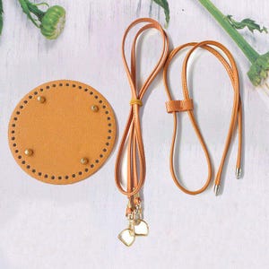 May include: A tan leather round base with pre-punched holes and gold-coloured rivets, alongside a matching leather strap with a gold-coloured clasp and silver-coloured tips. The strap is looped and tied with a yellow rubber band. The items are for crafting.