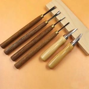 May include: A collection of six wood carving tools with different blade profiles. The handles are crafted from dark and light wood, featuring metal ferrules. The tools are displayed on a light orange surface, with a small wooden block in the background.