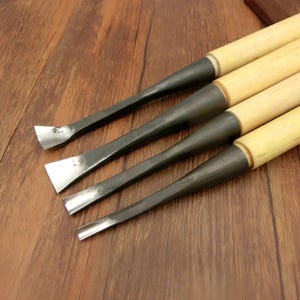 May include: Four wood carving chisels with wooden handles and dark metal blades. The blades have different shapes, including a flat edge, a V-shaped gouge, and a curved gouge. The tools are arranged diagonally on a wooden surface.