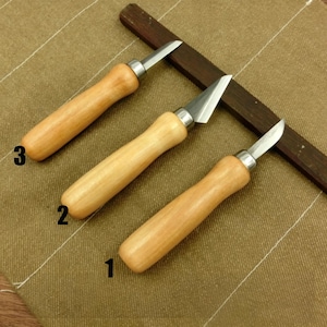 Puede incluir: Tres herramientas para tallar madera con mangos de madera marrón claro y cuchillas plateadas. Las cuchillas tienen diferentes formas: un borde recto, un borde en forma de V y un borde curvo. Una barra de madera marrón oscuro descansa detrás de las herramientas.