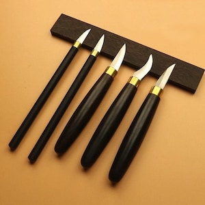 May include: Set of five wood carving tools with black handles and silver blades. The blades have a gold-colored band at the base. The tools are arranged on a dark brown wooden base, set against a light brown background.